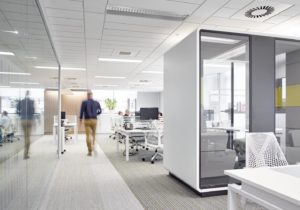 A light and airy office with a blurred figure walking through in blue shirt and yellow trousers. People work at computers in the background.